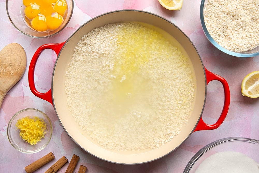 A pan filled with arborio rice, lemon zest and water. Around the pan is the ingredients to make Arroz Doce.