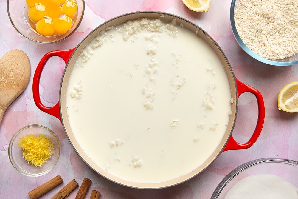 A pan filled with cooked arborio rice and milk. Around the pan is the ingredients to make Arroz Doce.
