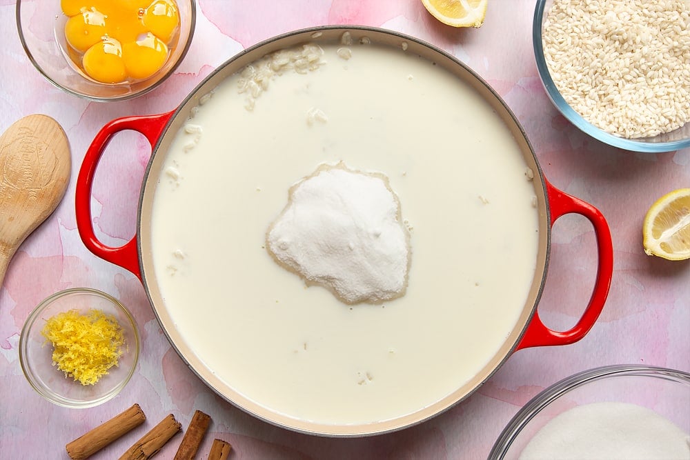 A pan filled with cooked arborio rice, milk and sugar. Around the pan is the ingredients to make Arroz Doce.