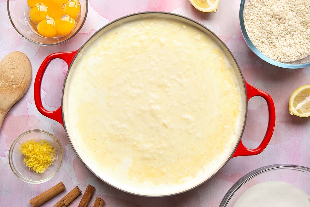 A pan filled with cooked arborio rice, milk, sugar and egg yolks. Around the pan is the ingredients to make Arroz Doce.