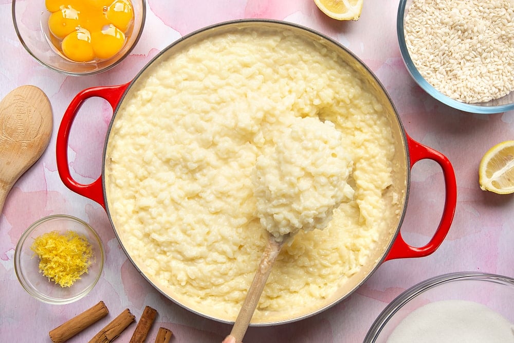 A pan filled with Arroz Doce. Ingredients to make the rice pudding surround the pan. A spoon lifts the thick pudding from the pan.