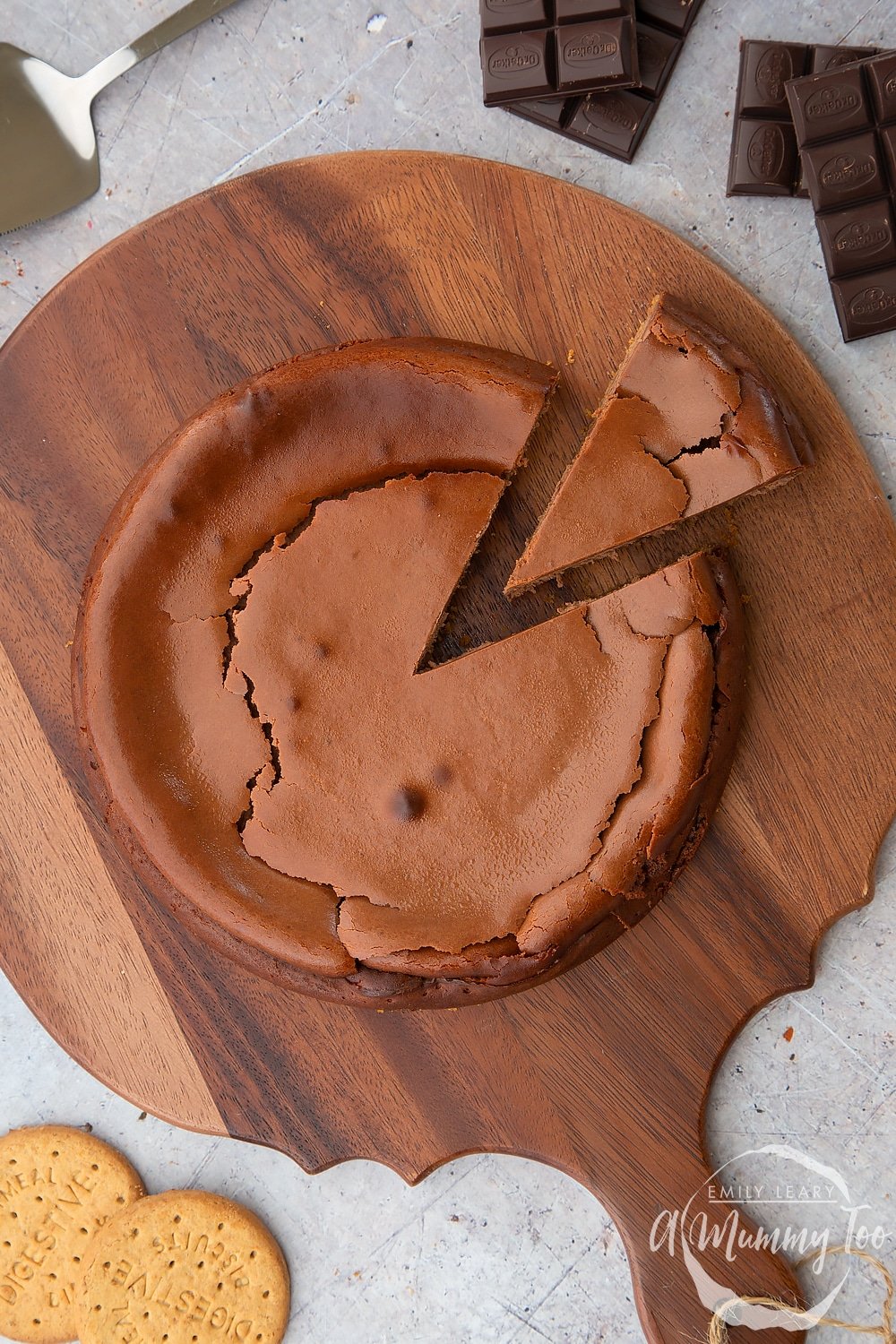 Overhead shot of the baked chocolate cheesecake with a slice already cut. The cheesecake sits on a wooden board surrounded by dark chocolate. 