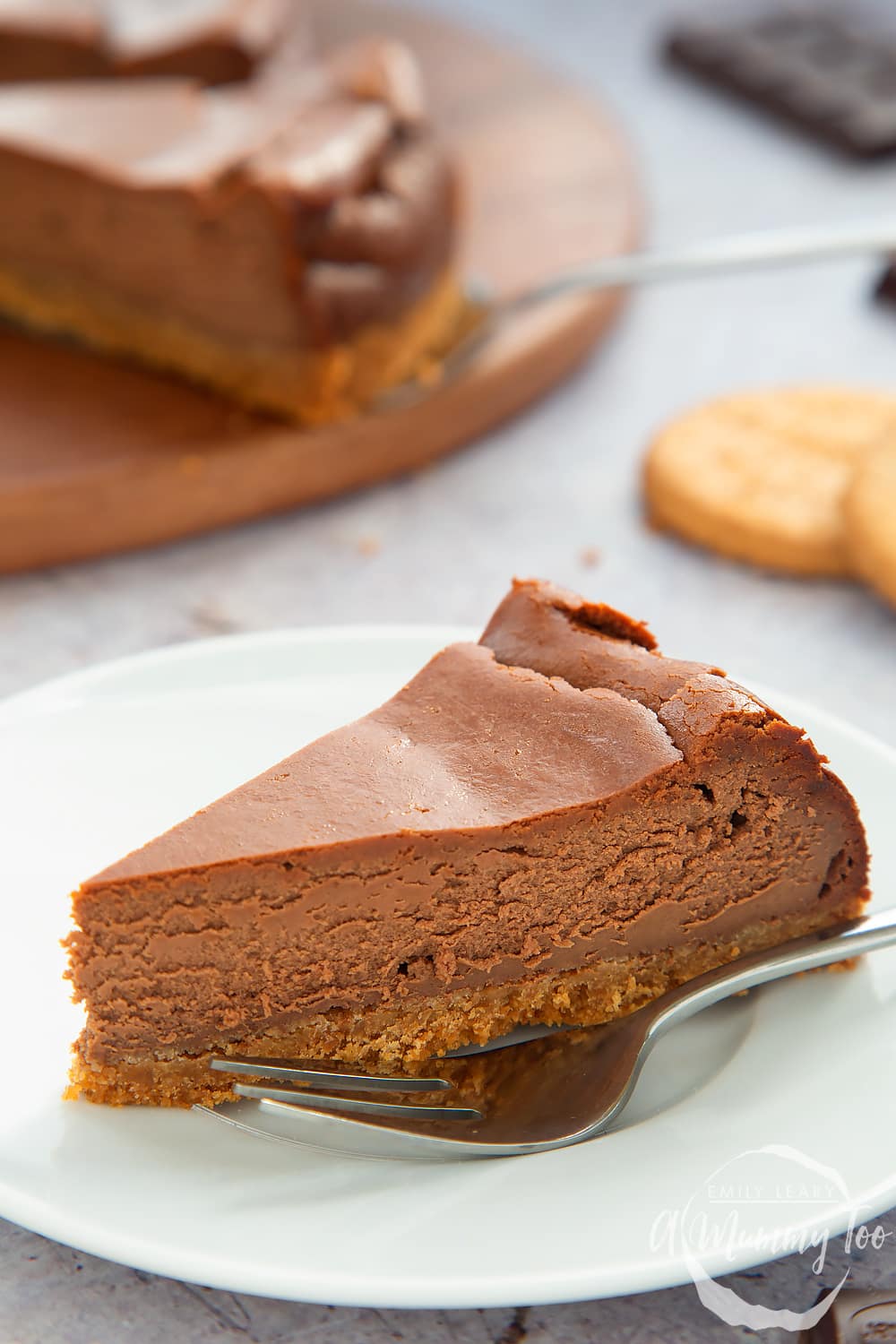 Close up side on view of a slice of the chocolate cheesecake on a white plate with a fork in the foreground. In the background you can see the additional slices of the baked chocolate cheesecake on a wooden board. 