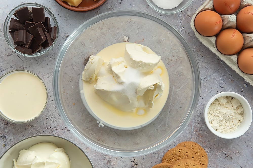 Overhead shot of the cream cheese, cream and sugar being mixed together in a pyrex bowl surrounded by some of the additional ingredients required to make the baked chocolate cheesecake. 