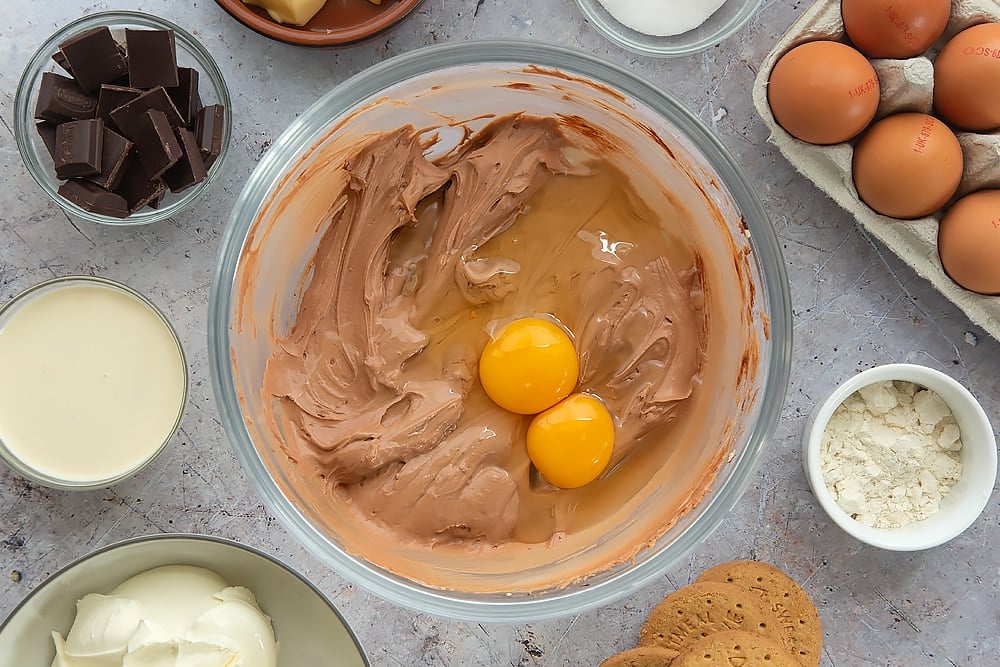 Adding two eggs to the baked chocolate cheesecake mixture. The overhead shot shows the bowl surrounded by some of the additional ingredients required for the baked chocolate cheesecake.