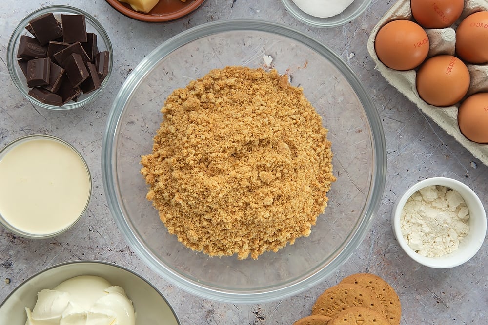 Overhead shot of the digestive biscuits required to make the baked chocolate cheese cake mashed up together inside a bowl surrounded by some of the additional ingredients required to make the baked chocolate cheesecake.