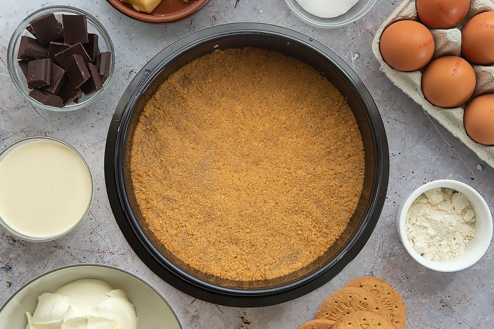 Overhead shot of the base of the cheesecake having been patted down until even and firm inside a tin and baked for five minutes. Some of the additional ingredients required for the baked chocolate cheesecake surround the tin. 