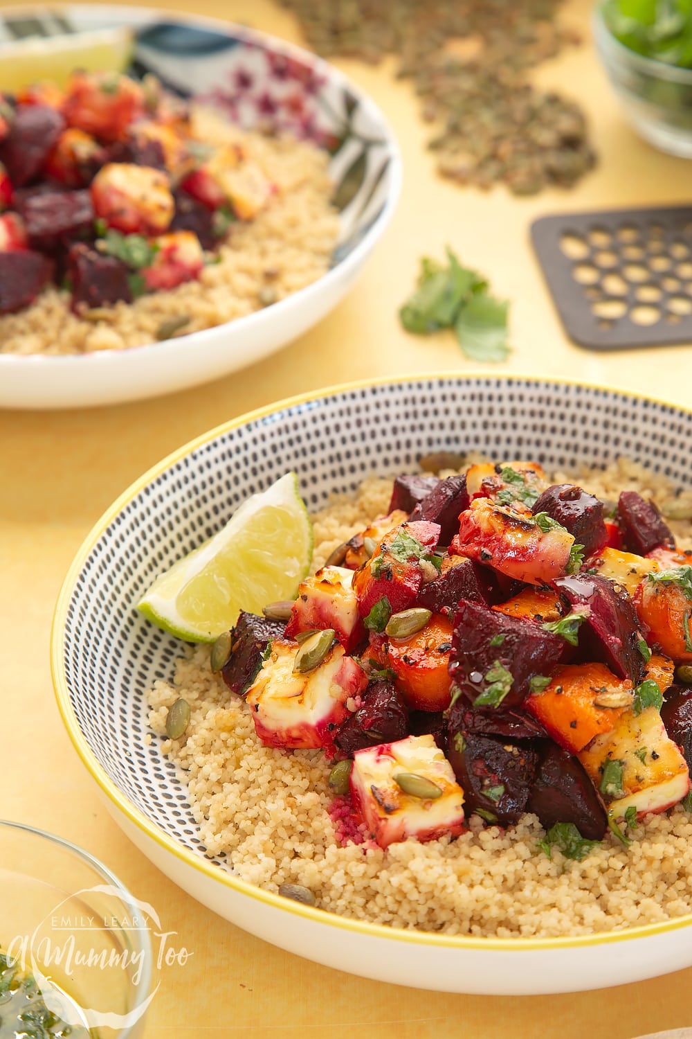 Side view of a yellow, black and white decoarive bowl filled with Beetroot and feta salad with couscous. In the background there is a floral bowl filled with Beetroot and feta salad with couscous and some ingredients required for the recipe.
