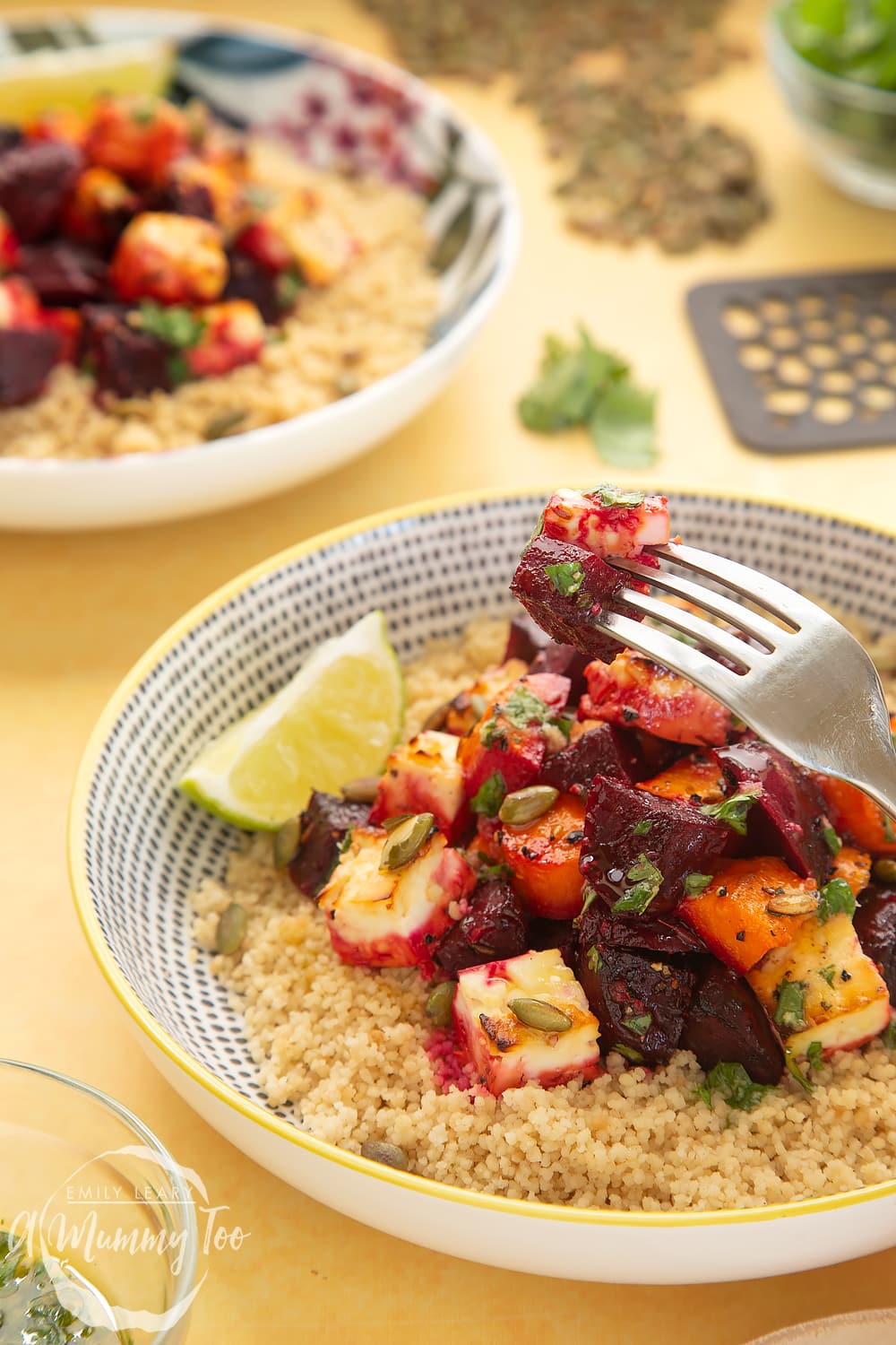 A side view of a decorative plate filled with Beetroot and feta salad with couscous ontop of a yellow table. In the background there's an additional bowl filled with Beetroot and feta salad with couscous and some of the ingredients used in the recipe. 