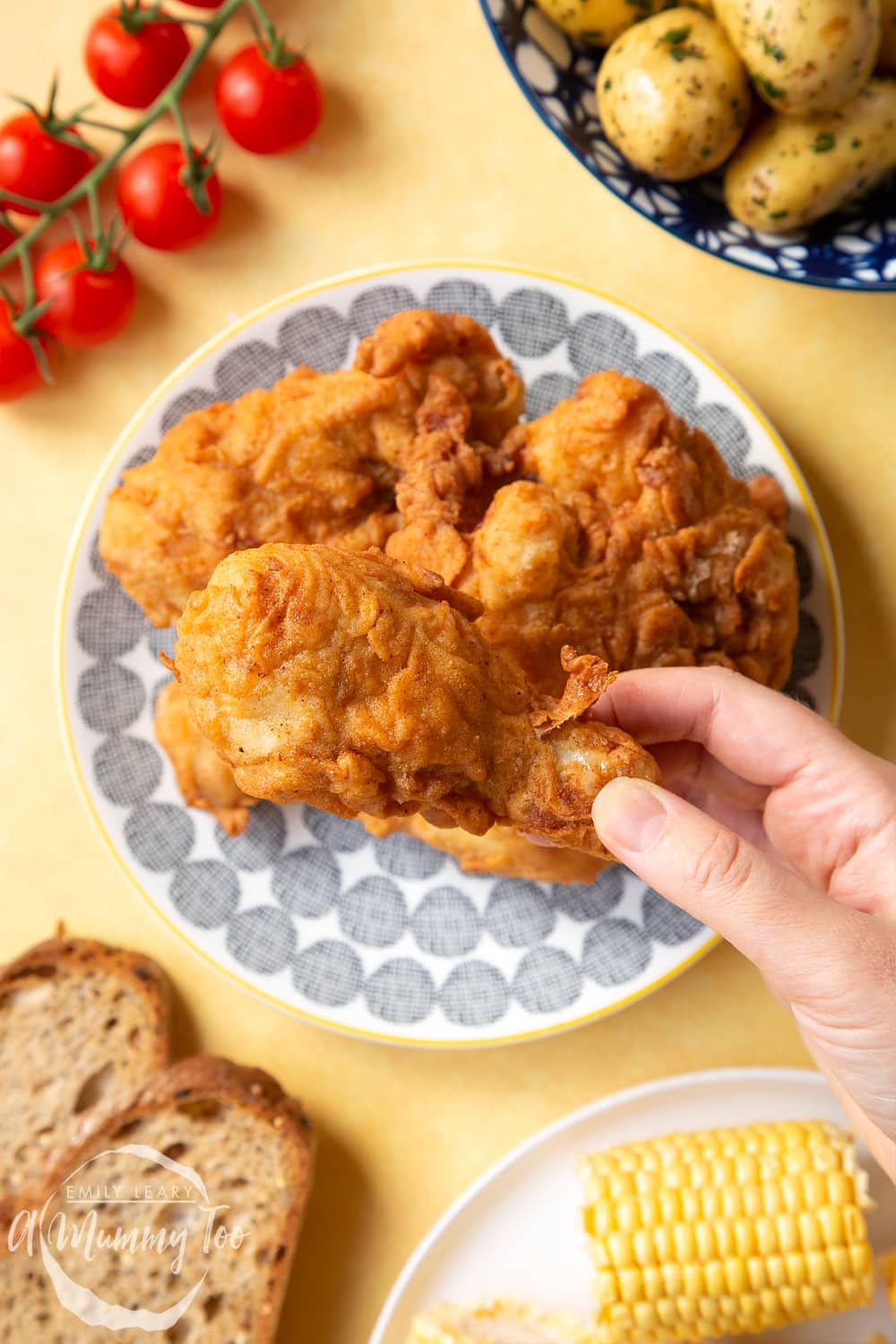 Gordon Ramsay’s buttermilk fried chicken arranged on a plate, surrounded by tomatoes, potatoes, bread and corn on the cob. A hand holds a well-coated drumstick, thickly coated with a seasoned, fried batter.