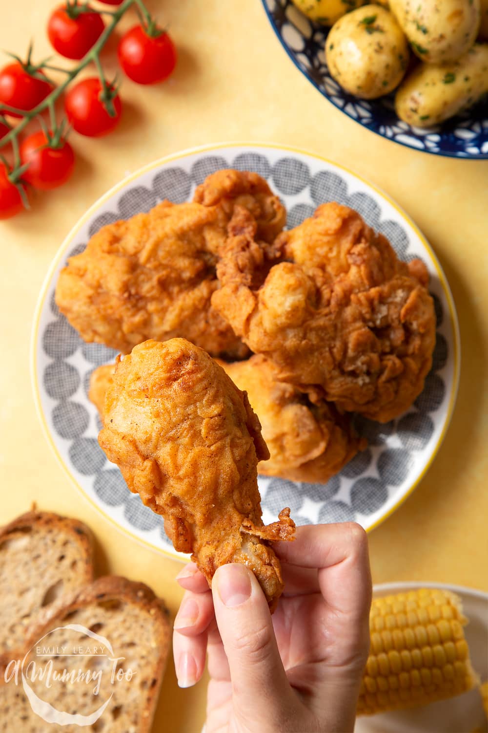 Gordon Ramsay’s buttermilk fried chicken arranged on a plate, surrounded by tomatoes, potatoes, bread and corn on the cob. A hand holds a well-coated drumstick, with a golden seasoned coating.