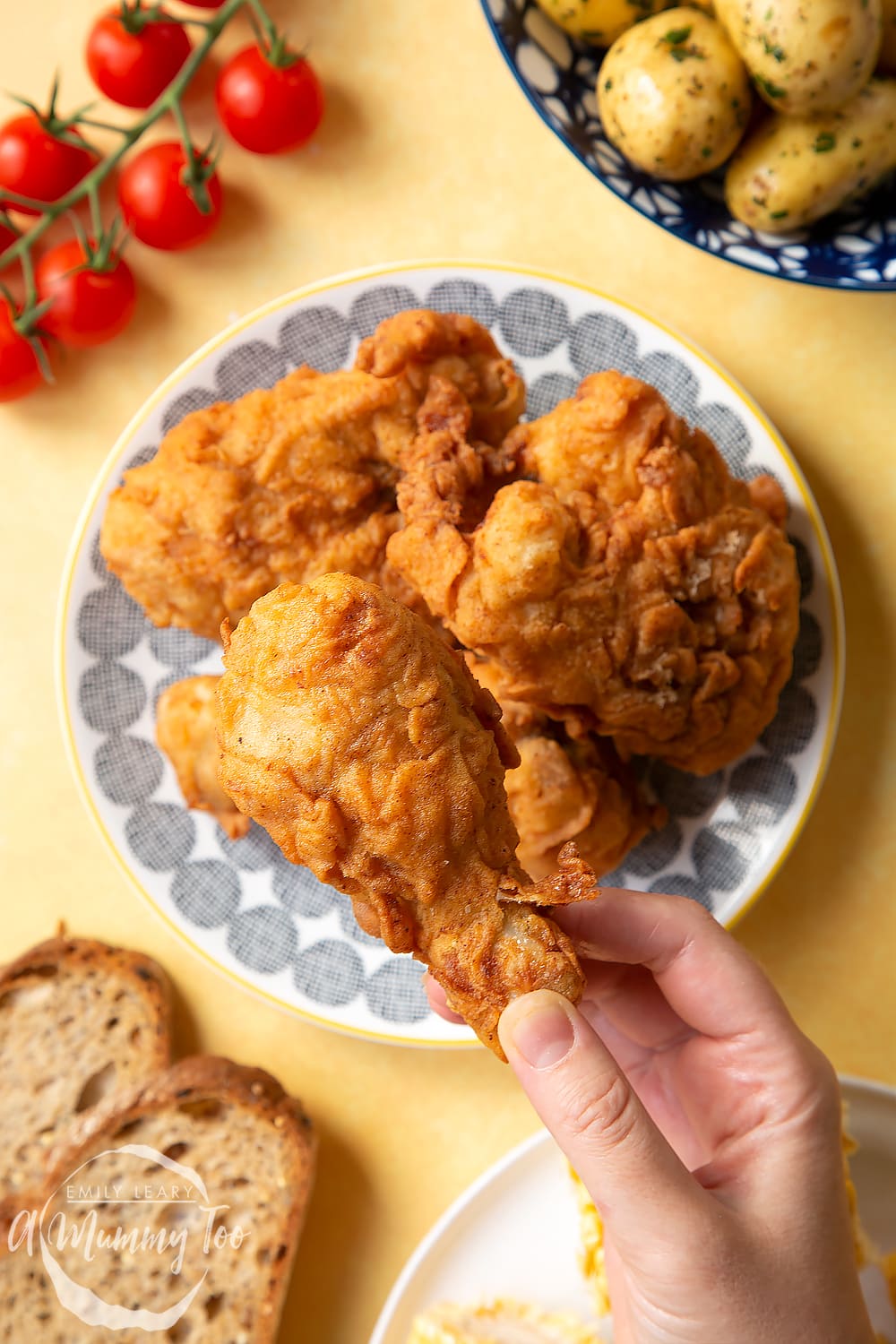 Gordon Ramsay’s buttermilk fried chicken arranged on a plate, surrounded by tomatoes, potatoes, bread and corn on the cob. A hand holds a well-coated drumstick.