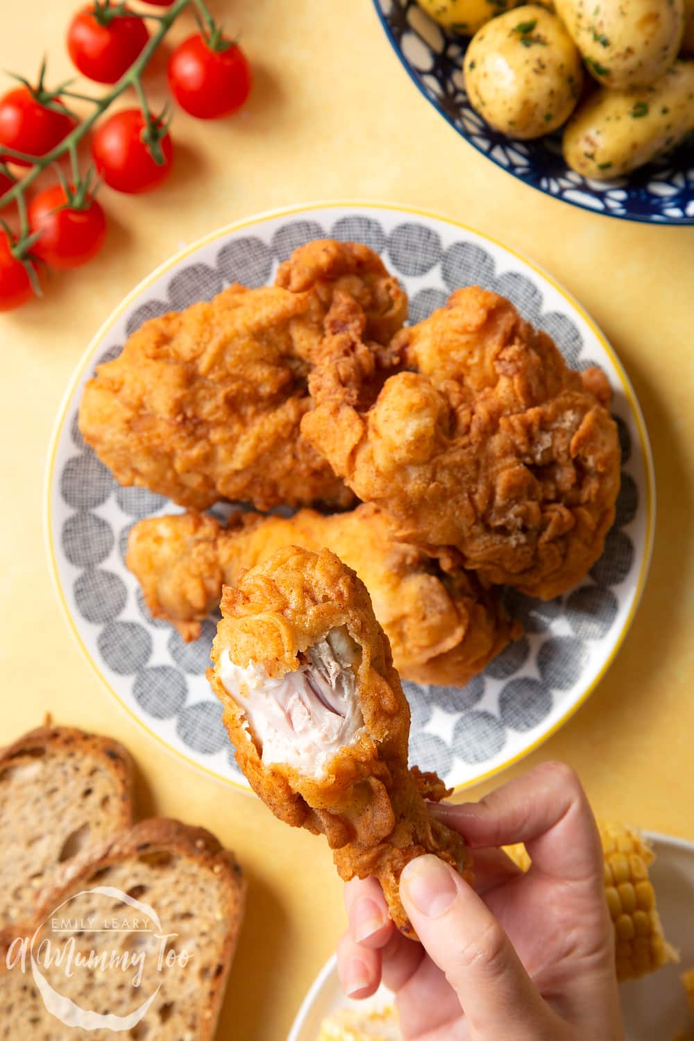 Gordon Ramsay’s buttermilk fried chicken arranged on a plate, surrounded by tomatoes, potatoes, bread and corn on the cob. A hand holds a well-coated drumstick, with the juicy chicken showing underneath the seasoned coating.