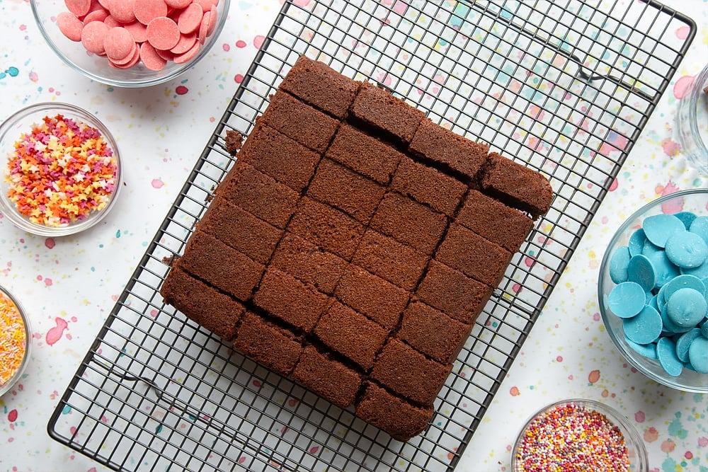 A freshly baked chocolate cake cooling on a wire rack, cut into pieces. Ingredients to make a cake pop bouquet surround the cake.