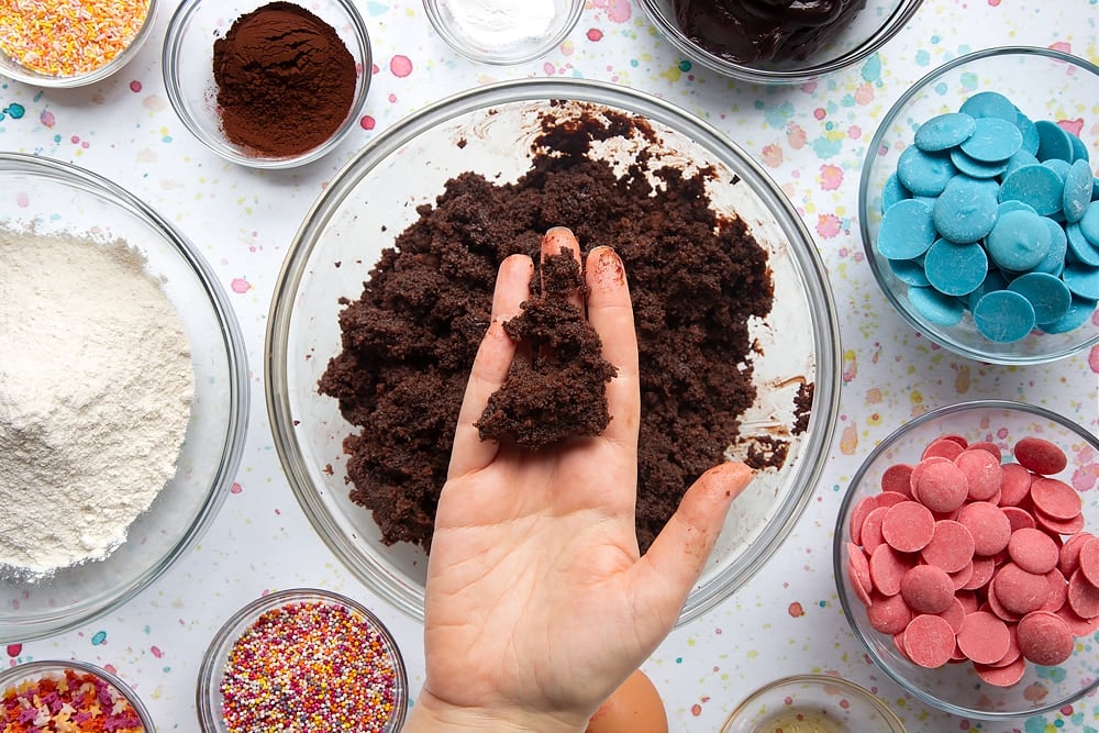 chocoladecake verkruimeld in een kom en gemengd met chocolade glazuur. Een hand houdt een stuk van het mengsel vast. Ingrediënten om een cake pop boeket te maken omringen de kom.