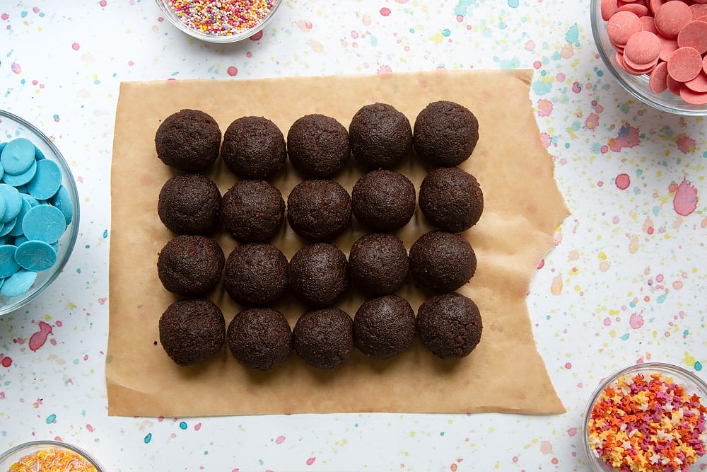 Rows of chocolate cake balls on baking paper. Ingredients to make a cake pop bouquet surround the paper.