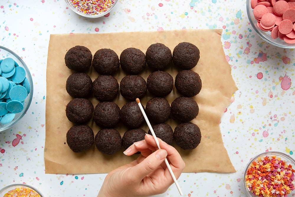 Rows of chocolate cake balls on baking paper. A hand holds a cake pop stick above them, dipped in chocolate. Ingredients to make a cake pop bouquet surround the paper.