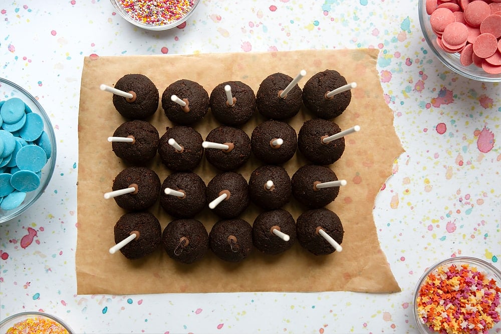 Rows of chocolate cake balls on baking paper. Cake pop sticks are pushed into each ball. Ingredients to make a cake pop bouquet surround the paper.