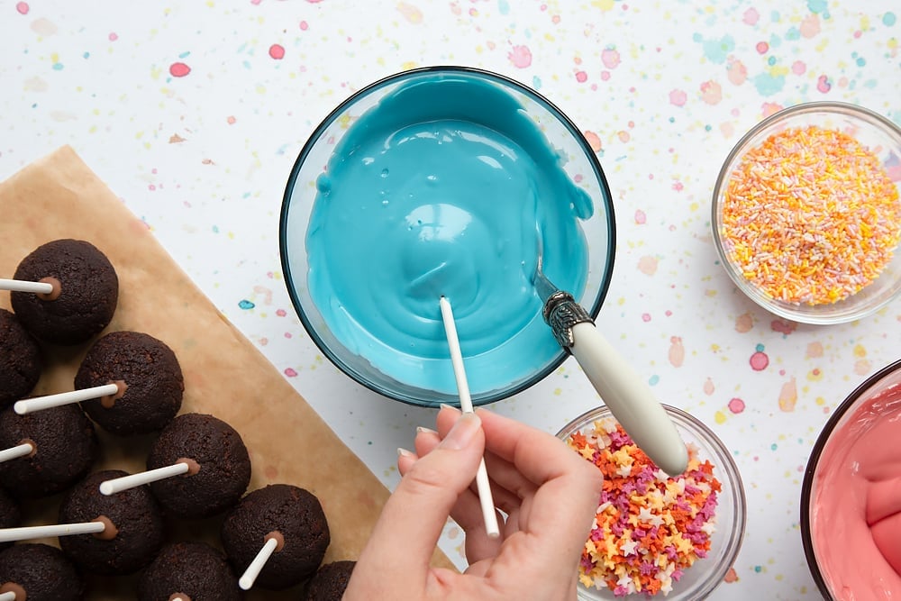 A bowl containing melted blue candy melts. A cake pop is being dipped into the bowl. Ingredients to make a cake pop bouquet surround the bowl.