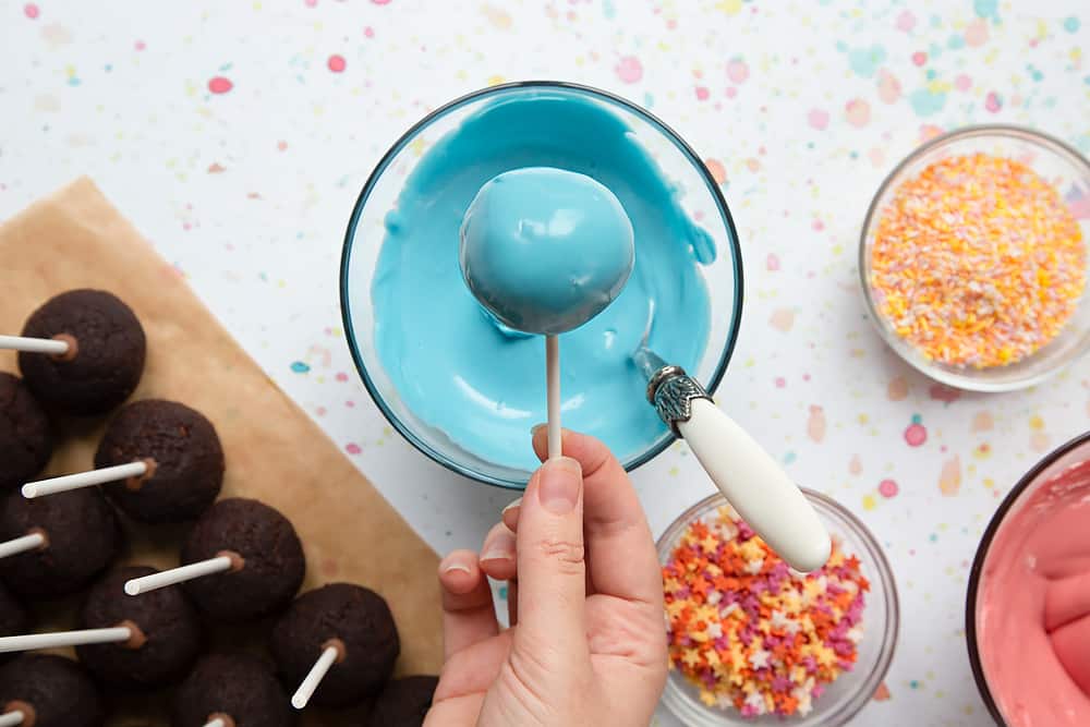 A bowl containing melted blue candy melts. A hand holds a freshly coated cake pop above the bowl. Ingredients to make a cake pop bouquet surround the bowl.