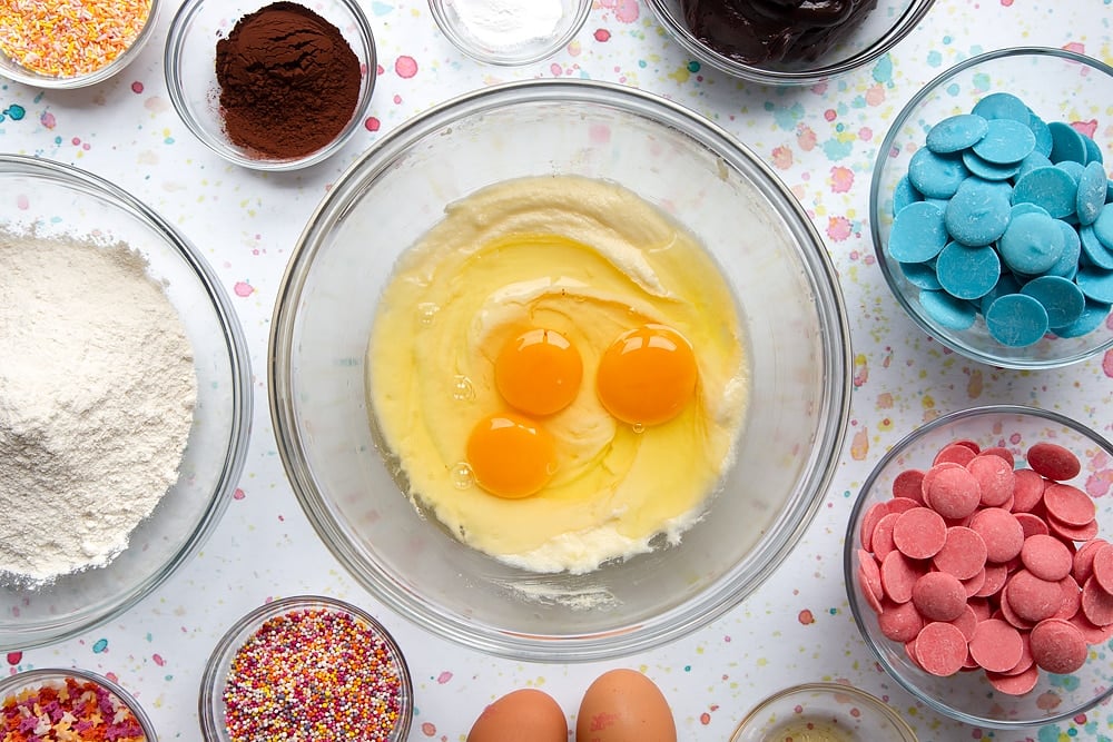 Azúcar y mantequilla en crema en un tazón con huevos por encima. Los ingredientes para hacer un ramo de pastel rodean el tazón.