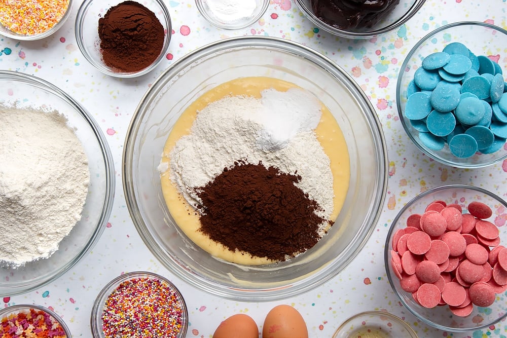  Zucchero, burro e uova mantecati insieme in una ciotola con farina, cacao e lievito in cima. Ingredienti per fare una torta pop bouquet circondano la ciotola.