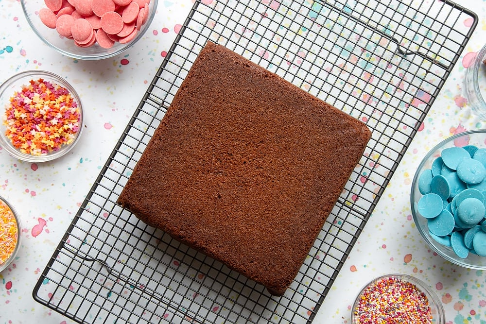 A freshly baked chocolate cake cooling on a wire rack. Ingredients to make a cake pop bouquet surround the cake.