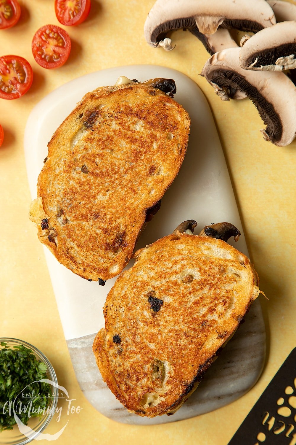 Two Jarlsberg cheese and mushroom toasties are on a serving board waiting to be cut in half and eaten.