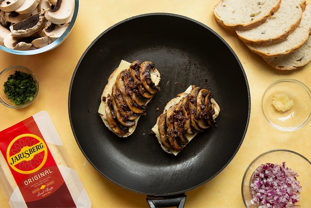 A frying pan with two slices of bread side by side and Jarlsberg cheese on top of both and carefully arranged cooked mushroom slices laid on top.