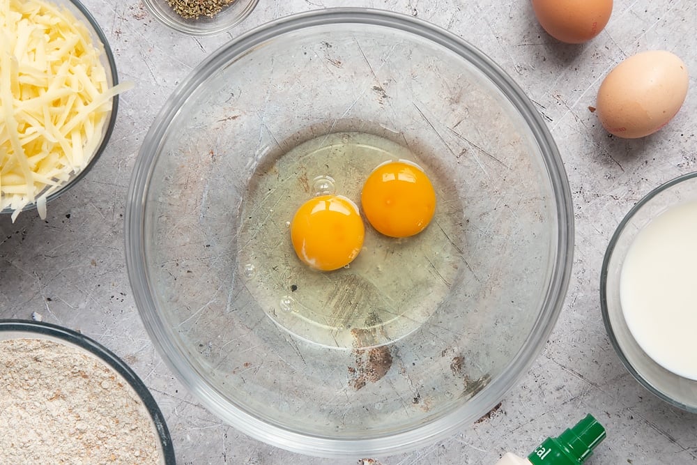 Eggs in a glass mixing bowl. Ingredients to make easy cheese muffins surround the bowl.