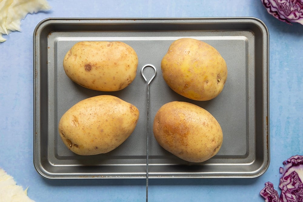 Four baking potatoes on a tray with a skewer. Ingredients to make jacket potato with coleslaw surround the tray.