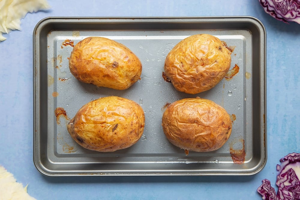 Four baked potatoes on a tray. Ingredients to make jacket potato with coleslaw surround the tray.