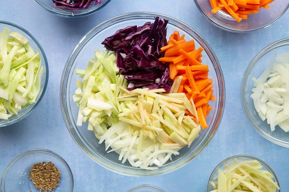 A glass mixing bowl containing carrot, onion, apple, white cabbage and red cabbage all cut into thin batons. Ingredients to make coleslaw without mayo surround the bowl.