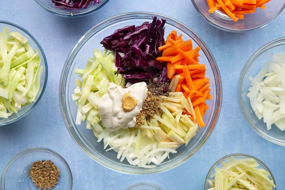 Red cabbage, white cabbage, carrot, onion, apple, crème fraiche, mustard and fennel seeds in a glass bowl. Ingredients to make jacket potato with coleslaw surround the bowl.