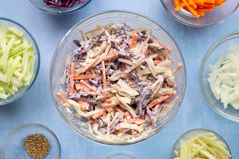 Red cabbage, white cabbage, carrot, onion, apple, crème fraiche, mustard and fennel seeds mixed together in a glass bowl. Ingredients to make jacket potato with coleslaw surround the bowl.