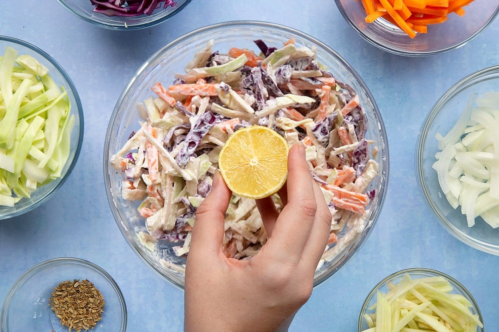 A glass mixing bowl containing shredded vegetables and creme fraiche. A hand is adding lemon juice. Ingredients to make coleslaw without mayo surround the bowl.