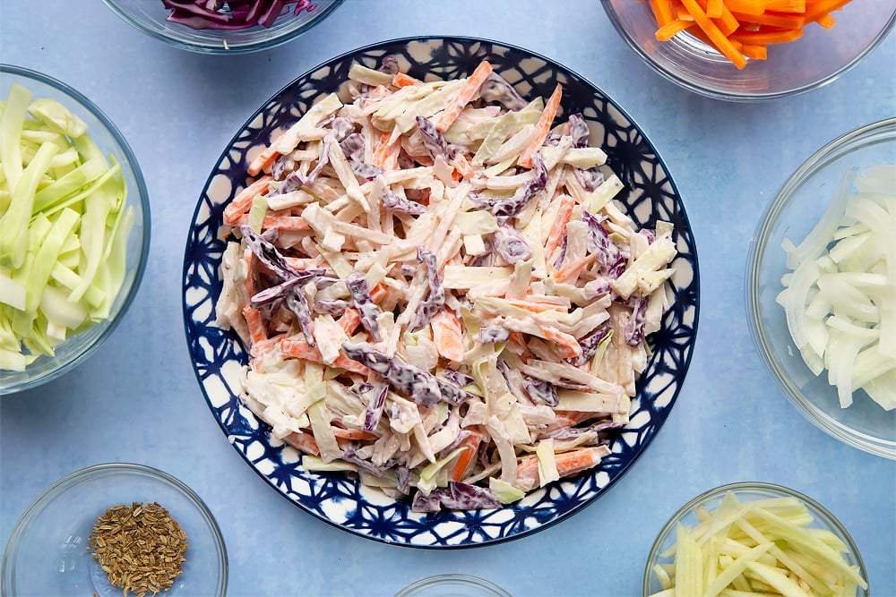 Coleslaw in a blue bowl. Ingredients to make jacket potato with coleslaw surround the bowl.