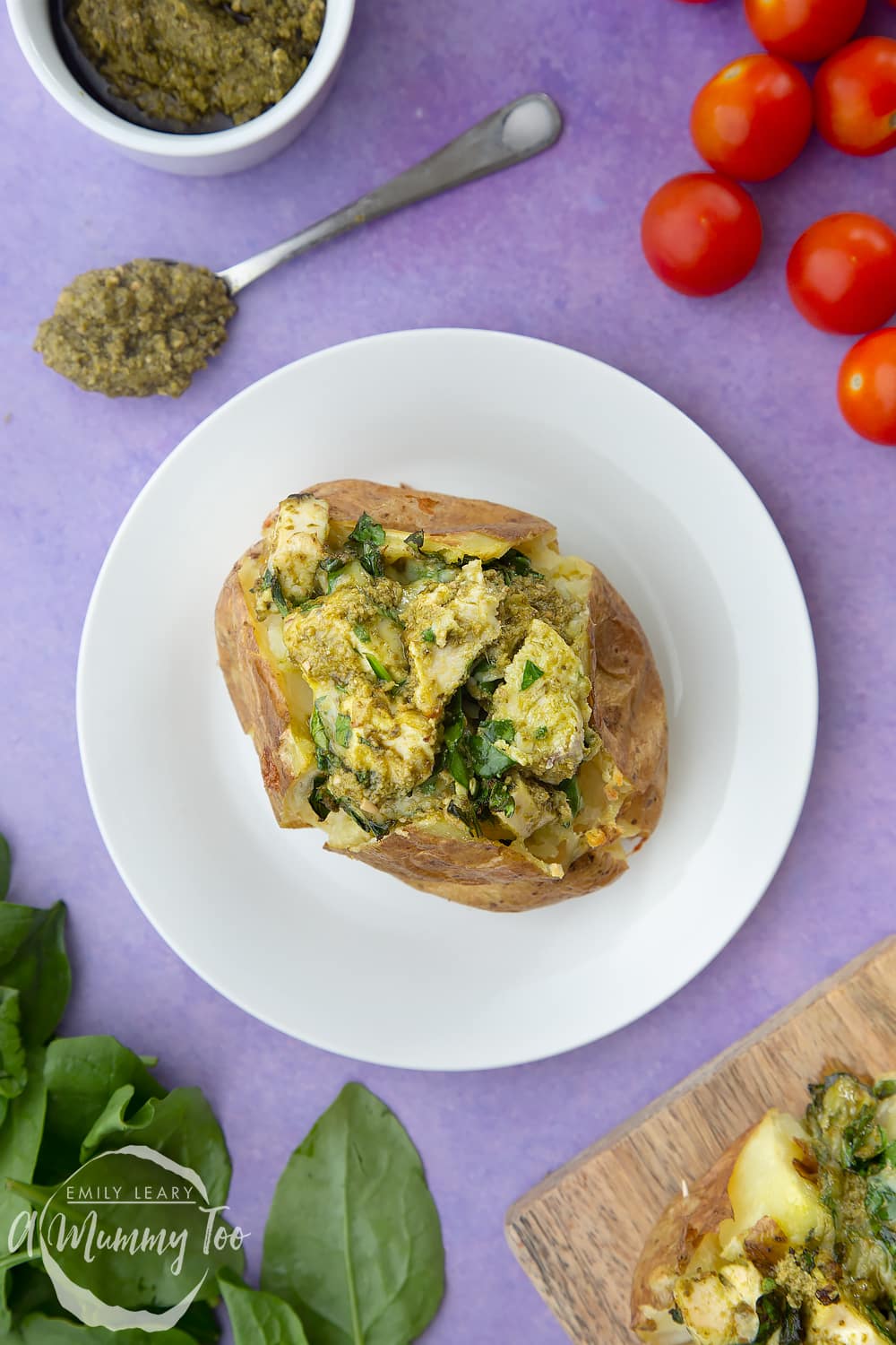 From above, a jacket potato with cheesy pesto chicken on a white plate. The potato is piled high with a pesto, spinach cheese and chicken mix. 