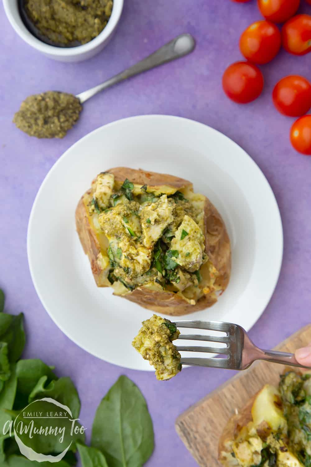 Jacket potato with cheesy pesto chicken on a white plate. The potato is piled high with a pesto, spinach cheese and chicken mix. A fork takes a bite.