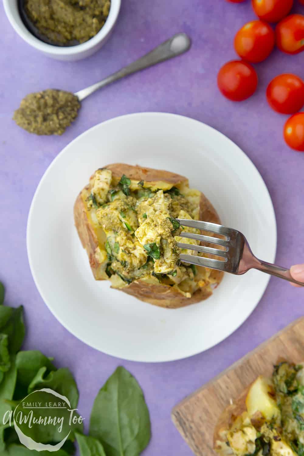From above, a jacket potato with cheesy pesto chicken on a white plate. The potato is piled high with a pesto, spinach cheese and chicken mix. A fork delves in.