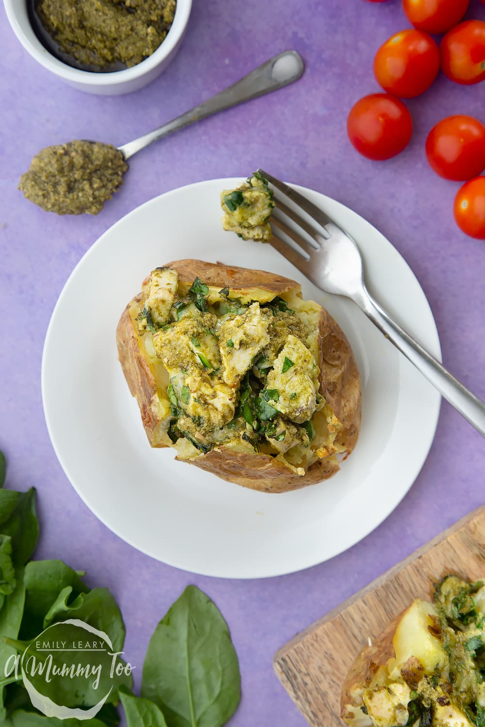 From above, a jacket potato with cheesy pesto chicken on a white plate. The potato is piled high with a pesto, spinach cheese and chicken mix. A fork rests to the side.