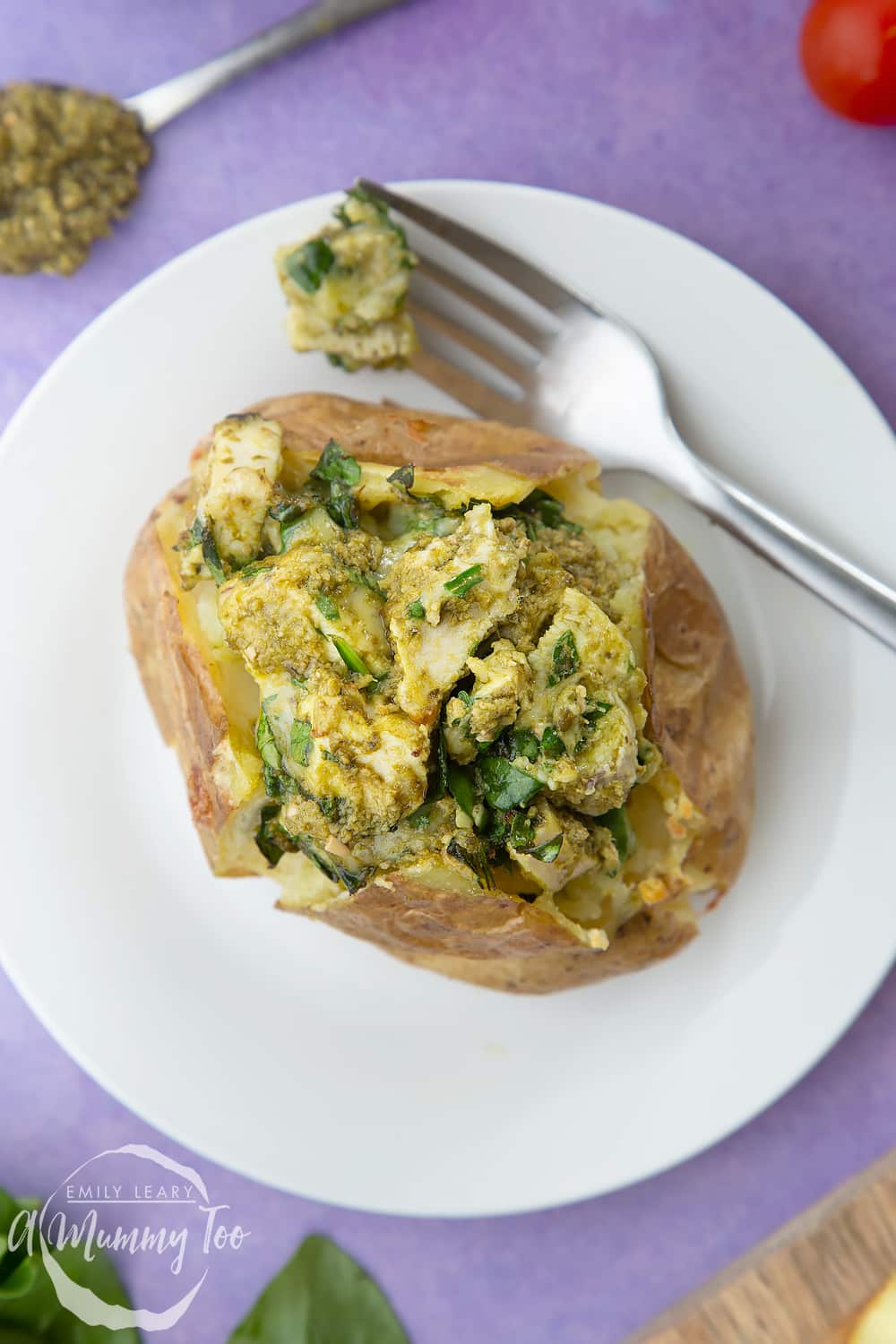 Jacket potato with cheesy pesto chicken on a white plate. The potato is piled high with a pesto, spinach cheese and chicken mix. A fork rests to the side.