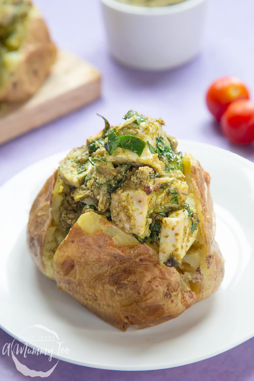 A jacket potato with cheesy pesto chicken on a white plate. The potato is piled high with a pesto, spinach cheese and chicken mix. More potatoes sit on a board in the background.