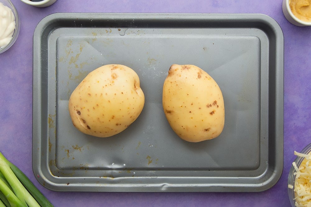 Raw baking potatoes on a baking tray. Ingredients to make jacket potato with cheesy pesto chicken surround the tray.