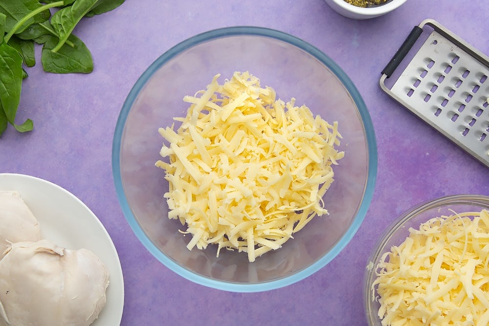Grated cheese in a mixing bowl. Ingredients to make jacket potato with cheesy pesto chicken surround the bowl.
