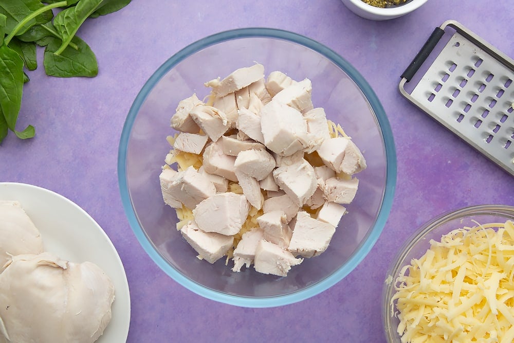 Grated cheese and chopped cooked chicken in a mixing bowl. Ingredients to make jacket potato with cheesy pesto chicken surround the bowl.