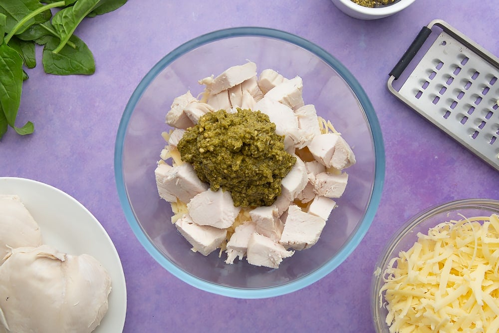 Grated cheese, chopped cooked chicken and pesto in a mixing bowl. Ingredients to make jacket potato with cheesy pesto chicken surround the bowl.