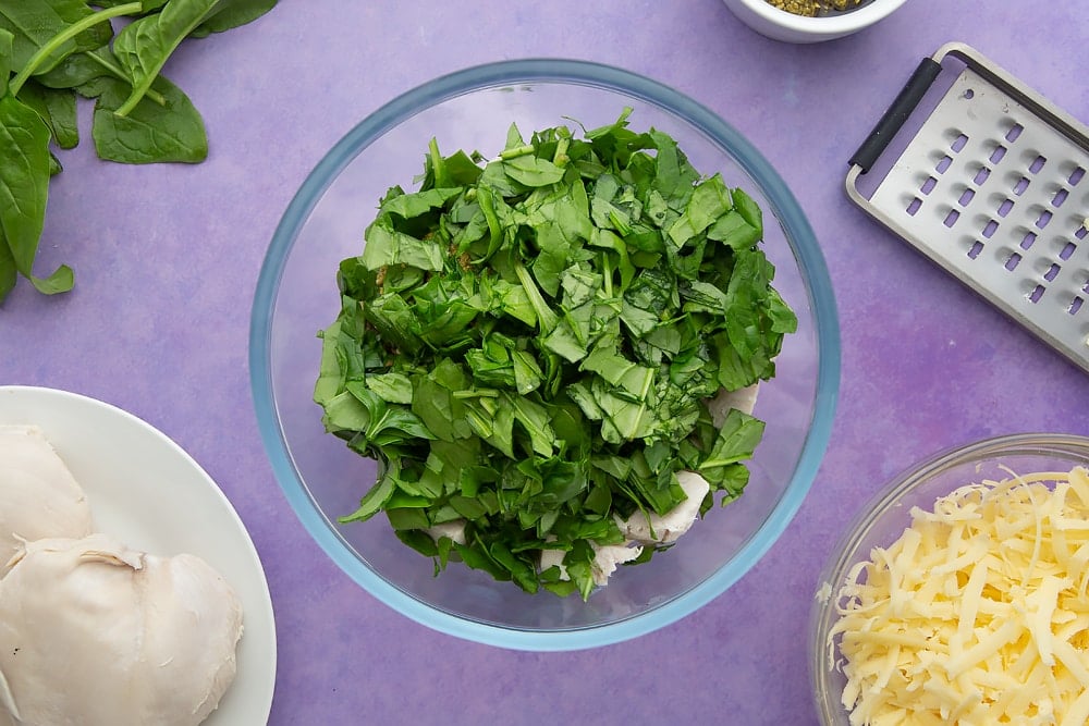 Grated cheese, chopped cooked chicken, pesto and fresh spinach in a mixing bowl. Ingredients to make jacket potato with cheesy pesto chicken surround the bowl.