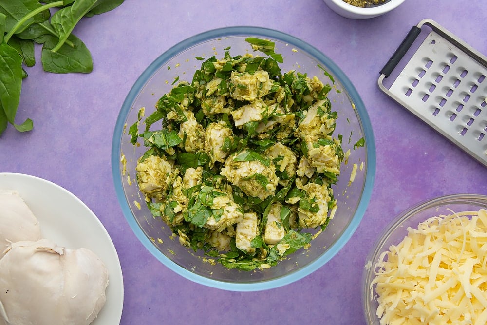 Cheesy pesto chicken in a mixing bowl. Ingredients to make jacket potato with cheesy pesto chicken surround the bowl.