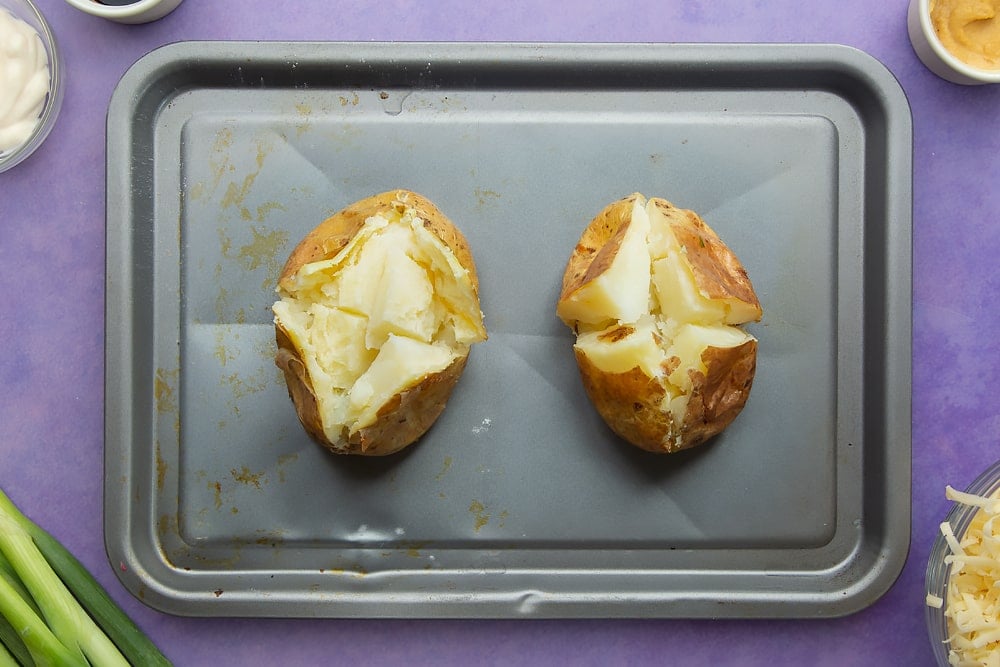 Baked potatoes cooked and cut open on a baking tray. Ingredients to make jacket potato with cheesy pesto chicken surround the tray.